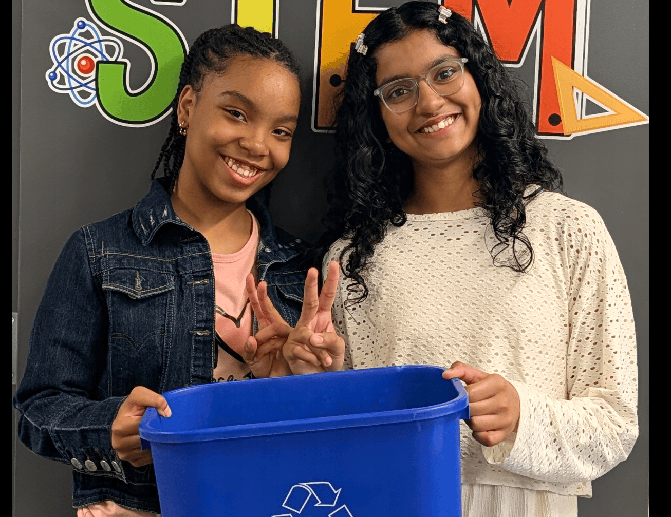Two teenage girls smile at the camera, they are both holding up peace signs and holding up a recycling bin