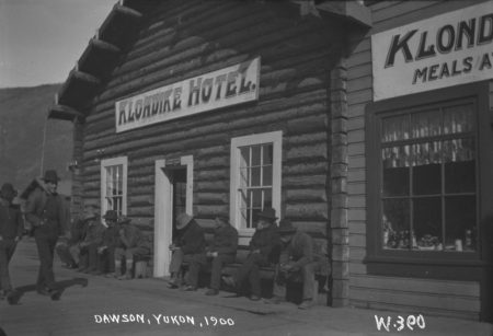 A black-and-white photo of a building with a sign reading "Klondike Hotel" and men sitting on a bench in front of it.