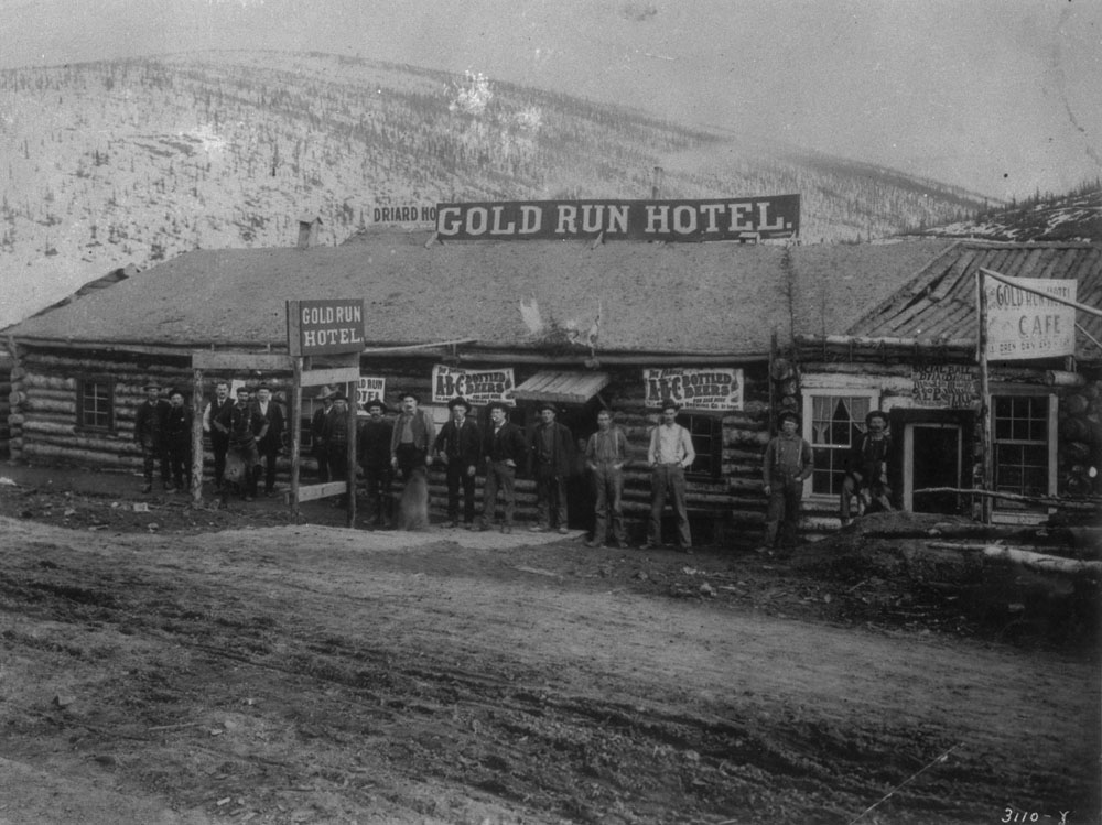 A black-and-white photo of a line-up of men standing outside a building in late 1800s Dawson City, Yukon, with a sign reading "Gold Run Hotel" on it.