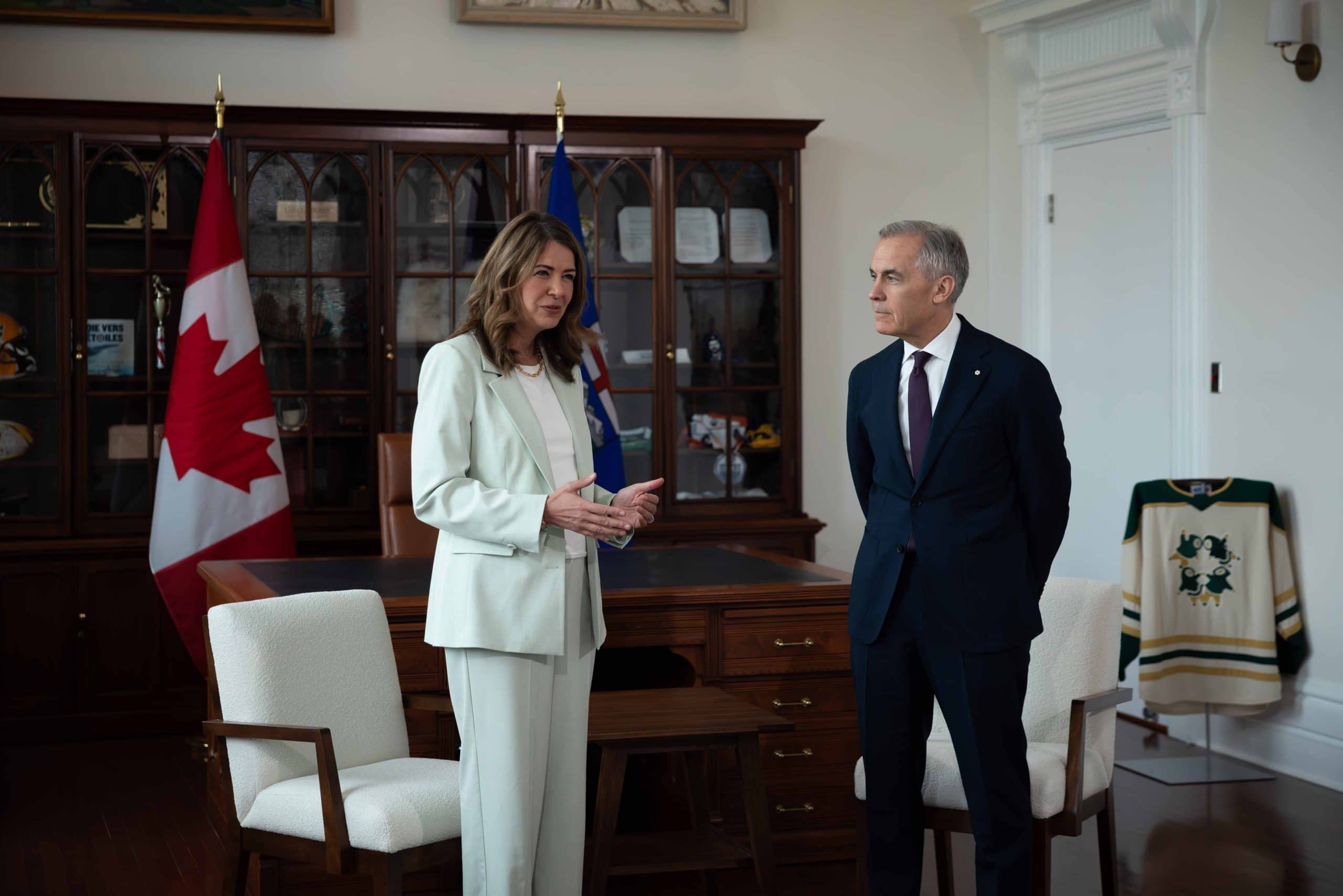 Woman in a white suit and man in a dark suit stand in an office with a Canada flag.
