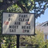 A letterboard sign reading "Big fans of clean air meet here, SAT 2 PM." IN the background is a big oak tree, blue skies and a house