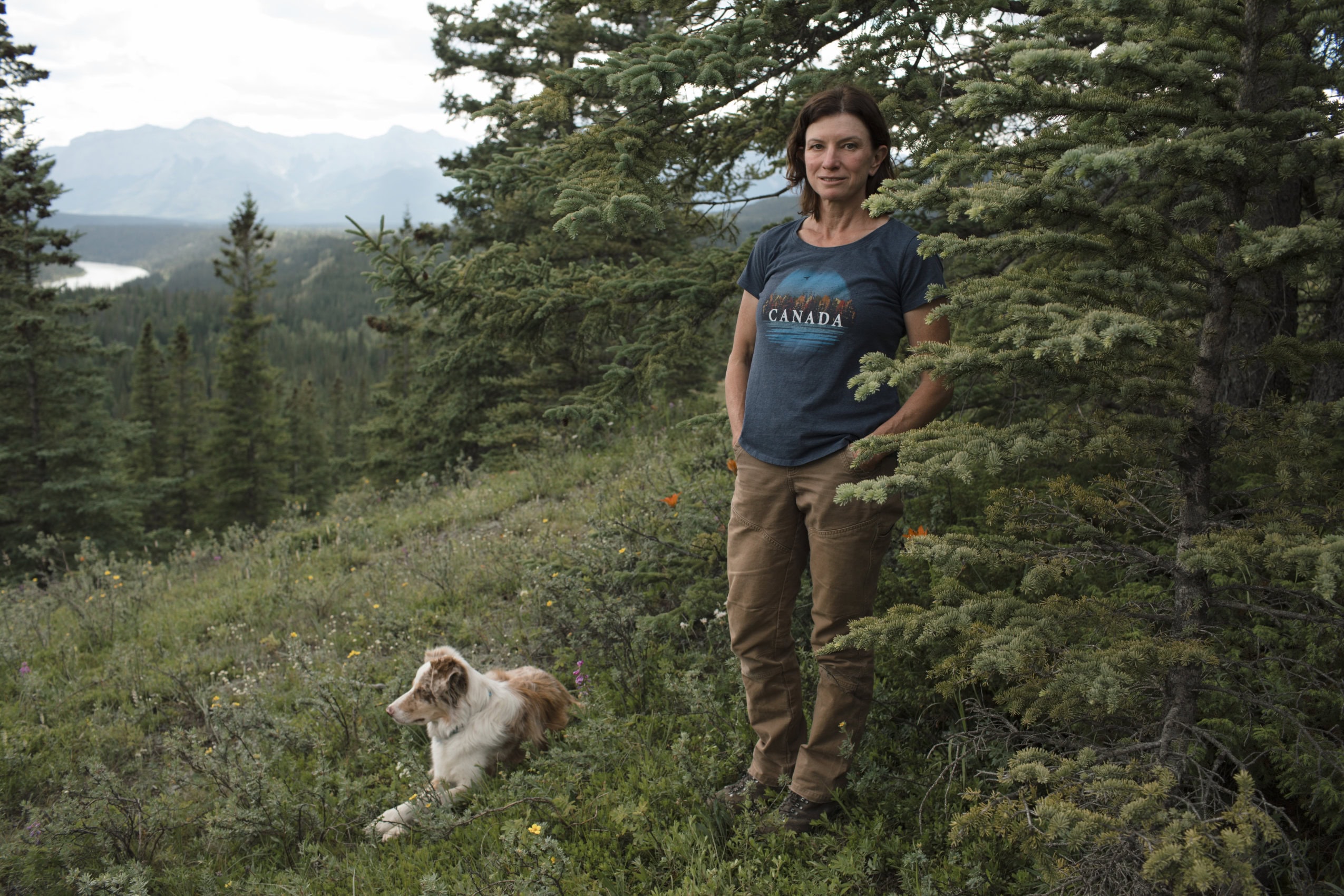 Michelle Wigmore stands on a grassy mountainside with trees at her back and her dog at her feet.