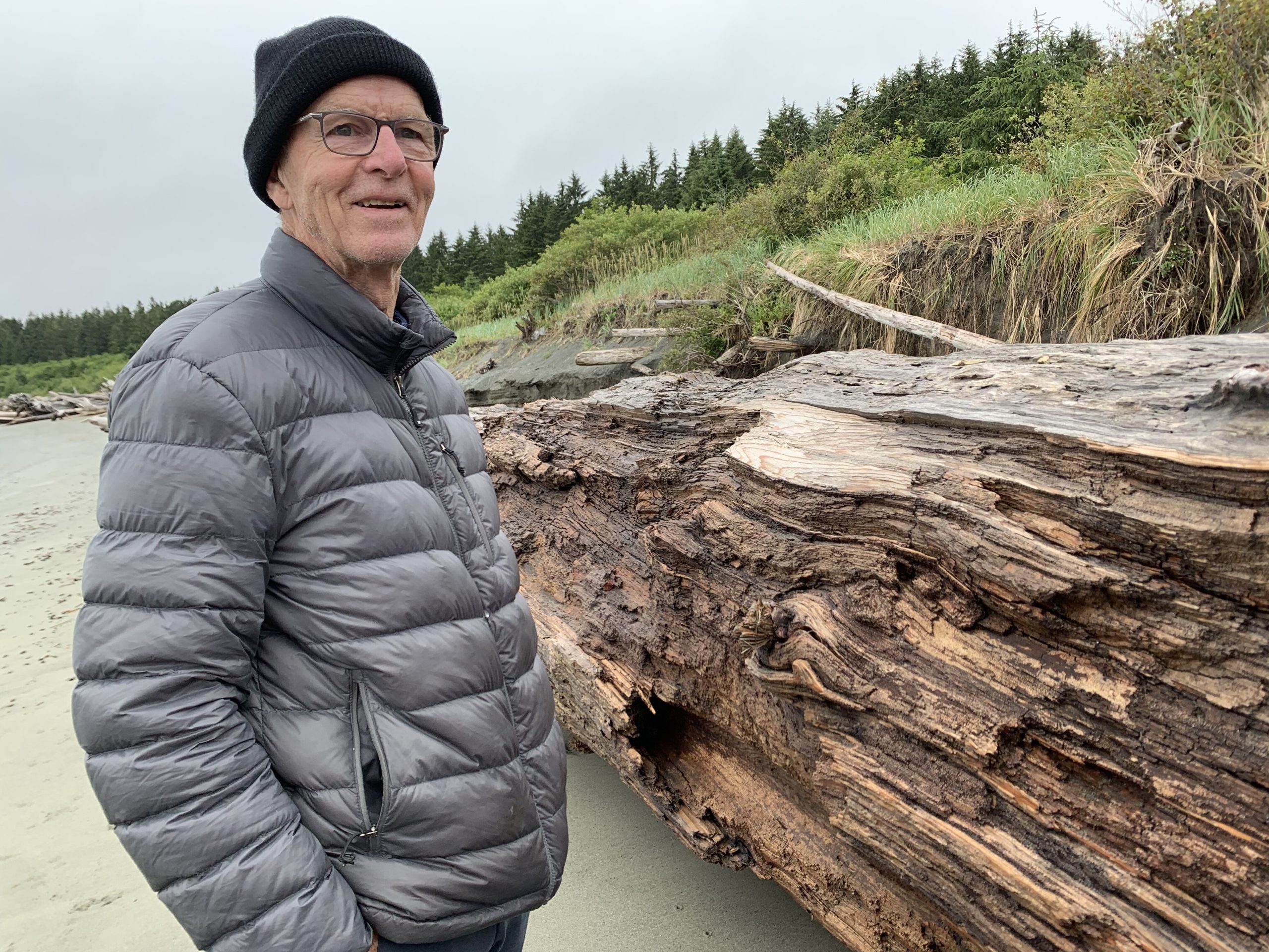 A man wearing a grey puffy jacket stands on a beach in front of a log 