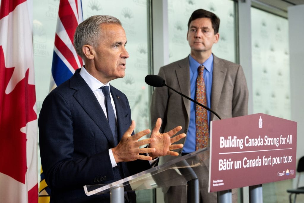 Prime Minister Mark Carney speaks into a microphone at a lectern while B.C. Premier David Eby looks on.