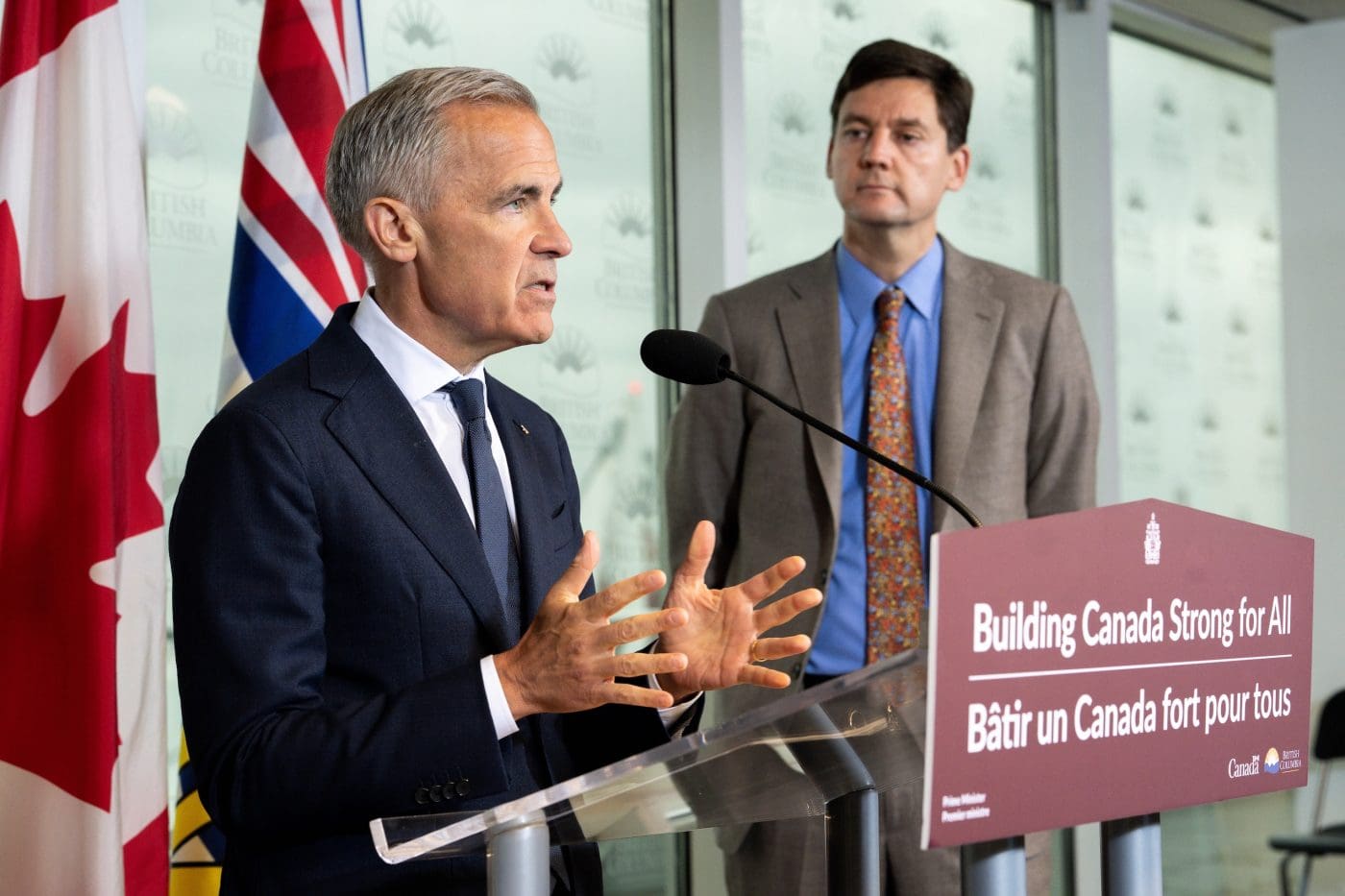Prime Minister Mark Carney speaks into a microphone at a lectern while B.C. Premier David Eby looks on.