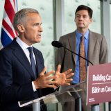 Prime Minister Mark Carney speaks into a microphone at a lectern while B.C. Premier David Eby looks on.