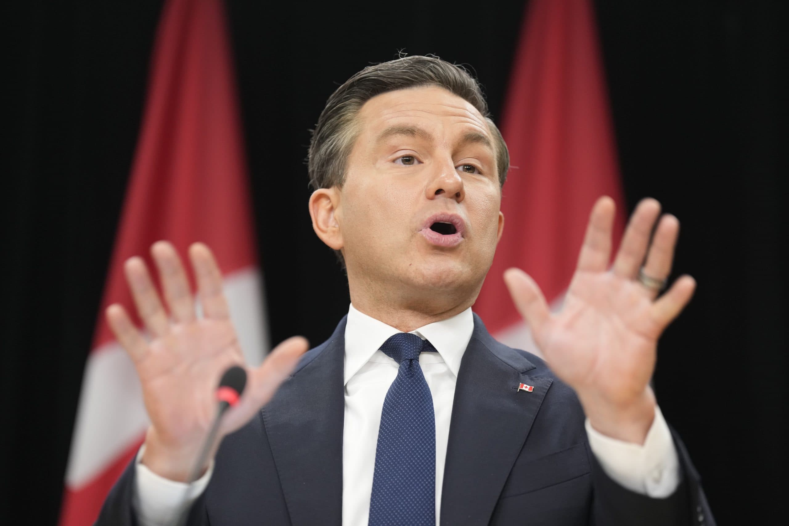 Man in a suit puts his hands up in a gesture while speaking into a microphone with Canadian flags in the background.