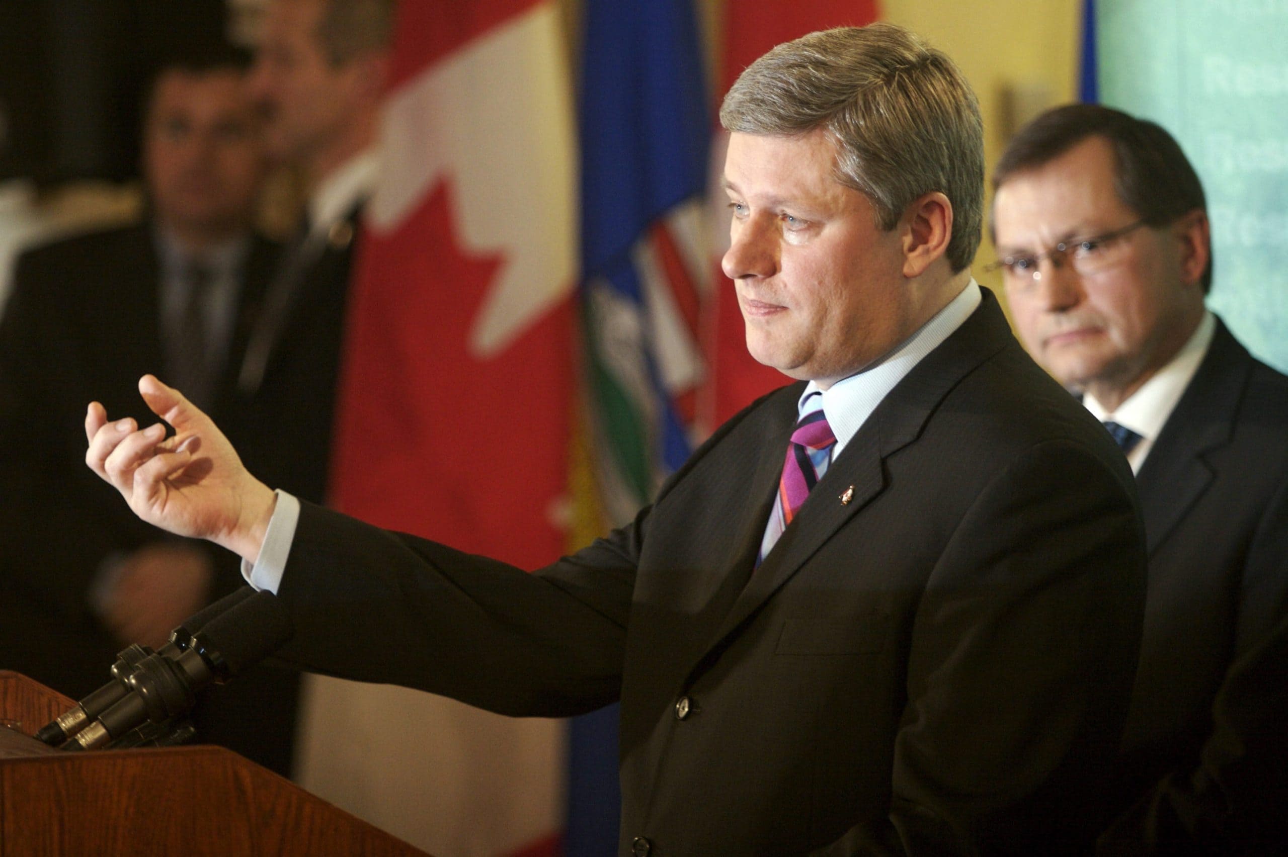Man in a dark suit speaks in front of a lectern while gesturing with his hand, as another man in a dark suit looks on