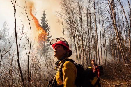 Two firefighters in gear look toward the sky as a fire burns in the forest behind them.