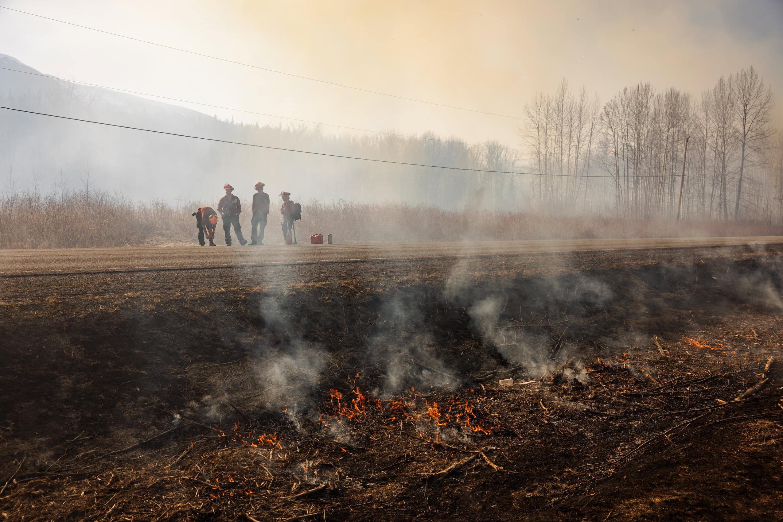 Four wildland firefighters stand on the side of a road while fire smoulders in the forest next to them.