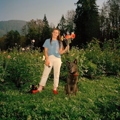 Alana Paterson with her dog in a field with a scenic background, holding a bouquet of bright orange flowers.