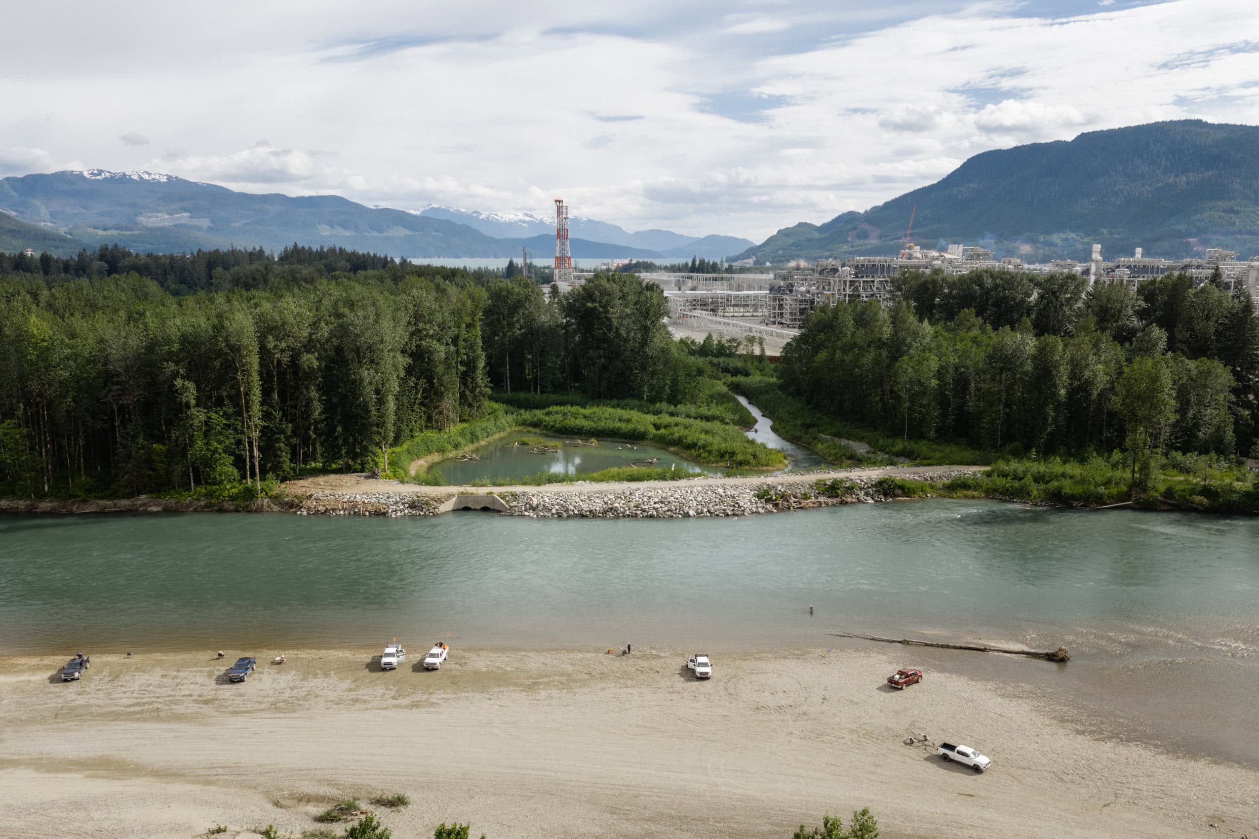 In the foreground, about a half dozen vehicles are parked near the shore of a river. In the background, LNG Canada's gas export facility in Kitimat, B.C.