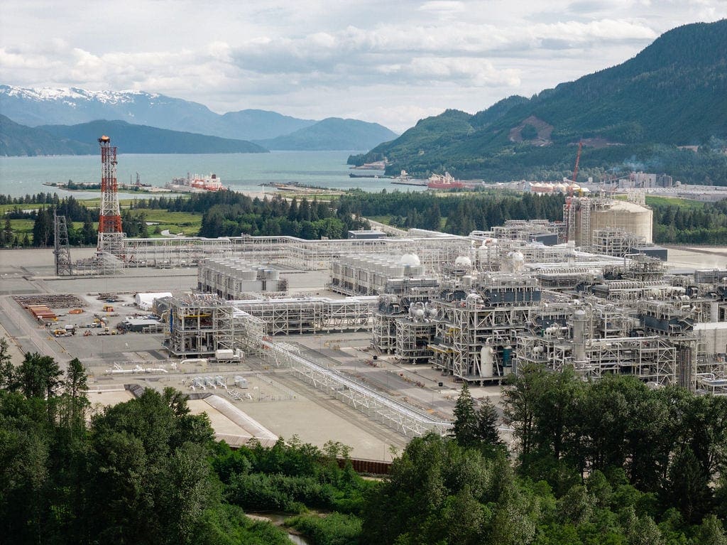 LNG Canada, a large industrial facility sits in the middle of the frame surrounded by green grass a river and mountains.
