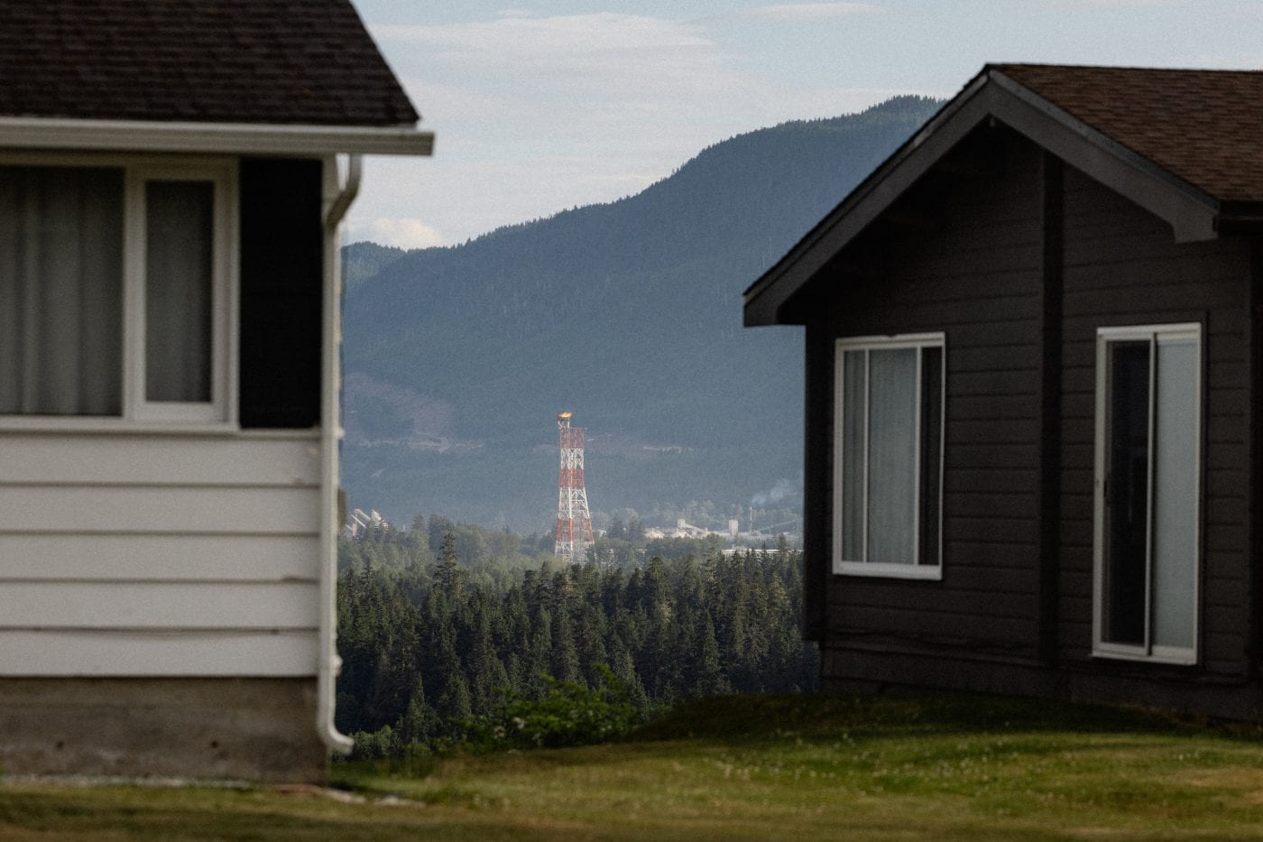 An LNG flare stack is seen in the distance, framed by two houses on either in the foreground