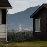 An LNG flare stack is seen in the distance, framed by two houses on either in the foreground