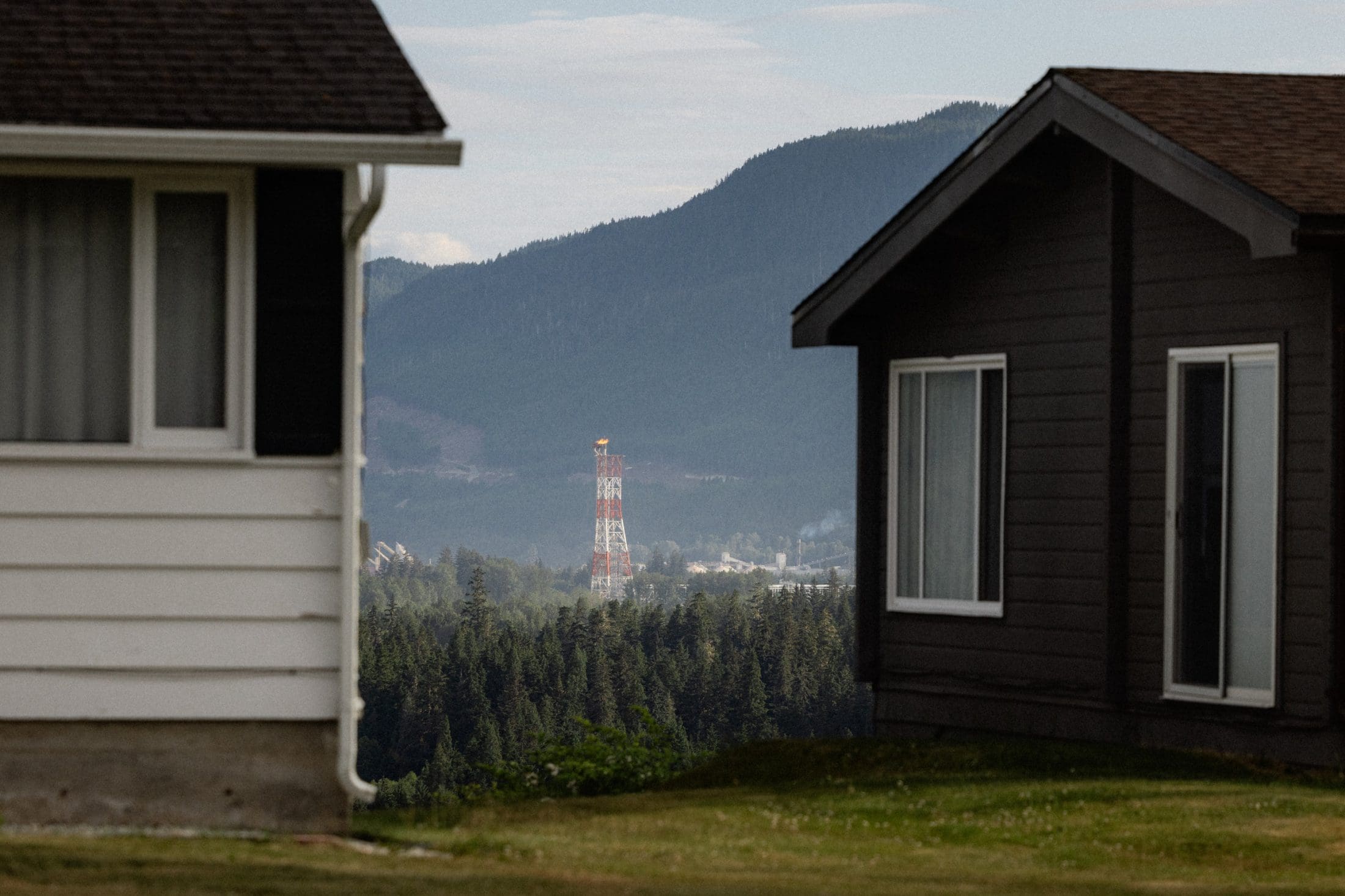 An LNG flare stack is seen in the distance, framed by two houses on either in the foreground