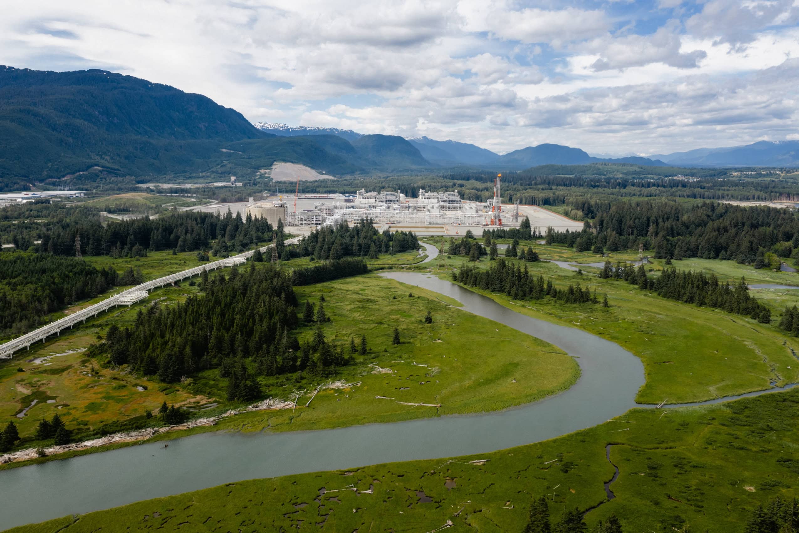 A river winds with green grass on either side until it meets a large industrial terminal.