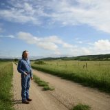 A man stands with one hand in his pocket on a dirt road that extends behind him through a green rural field.