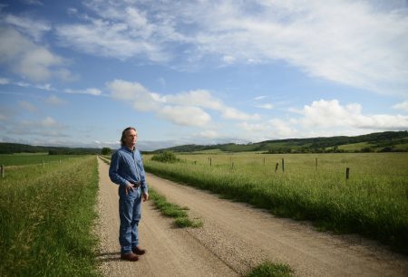 A man stands with one hand in his pocket on a dirt road that extends behind him through a green rural field.