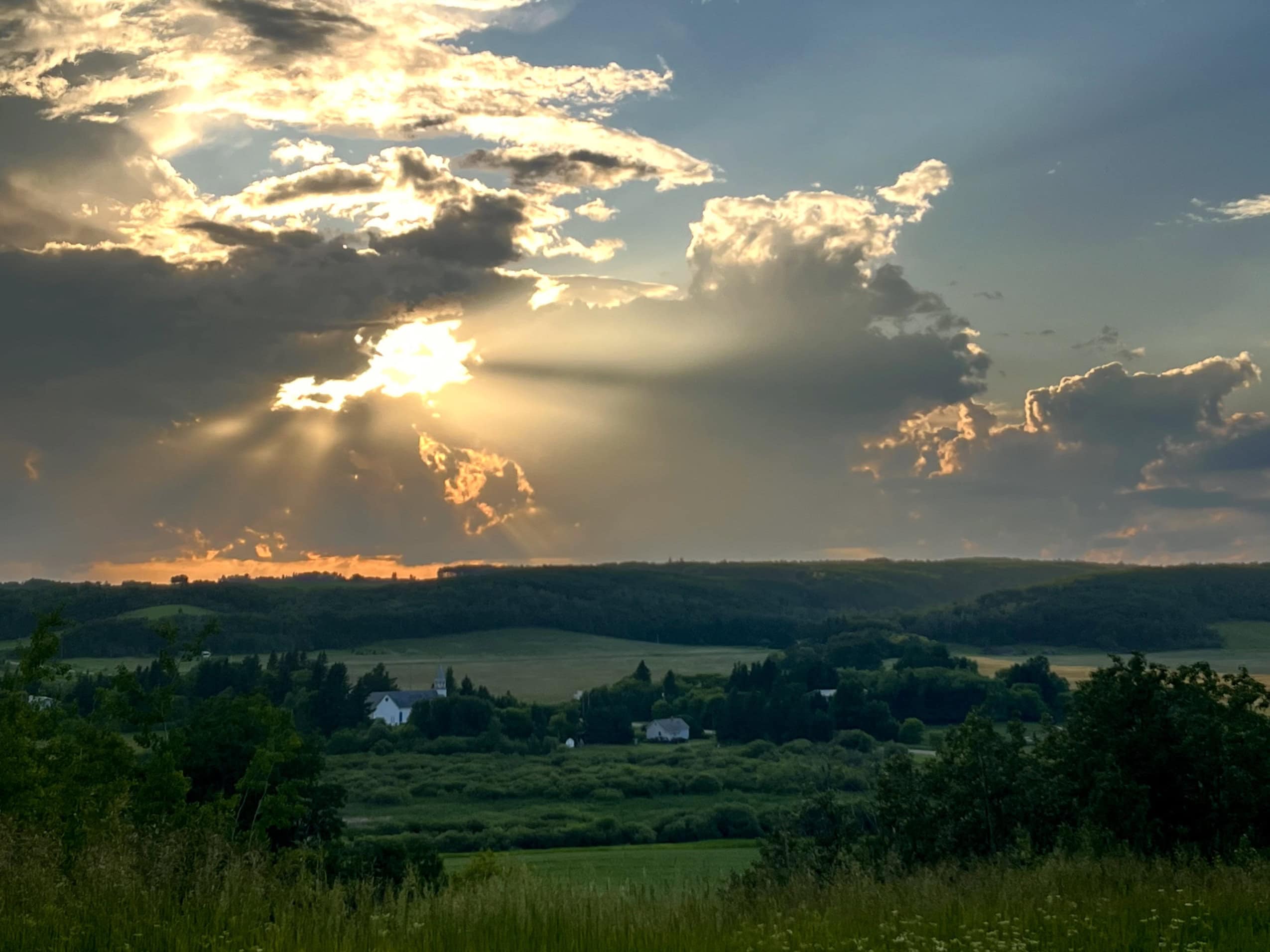 The sun, hanging low in the sky, pokes through clouds and sends rays of golden light streaming toward a rural community in Manitoba, nestled among lush summer fields.