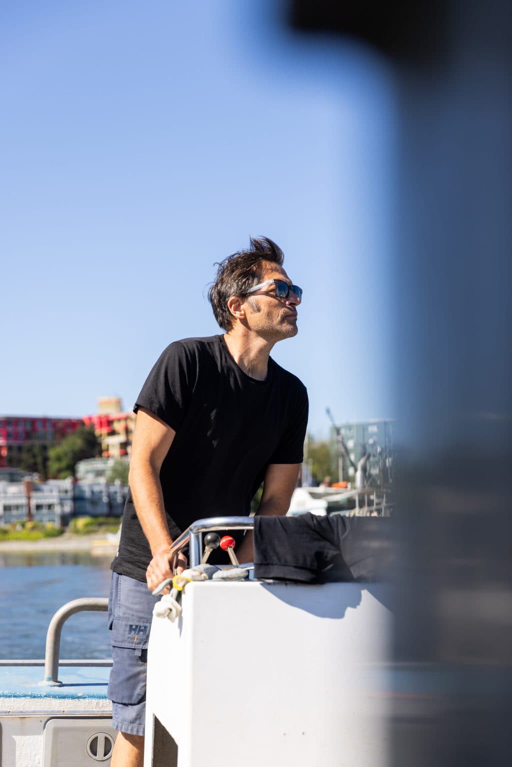 A man in a black t-shirt at the wheel of a boat on a sunny day.