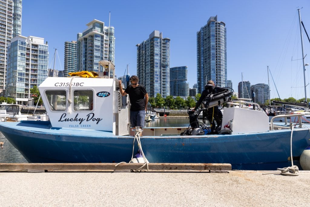 A man in a black t-shirt stands on a blue and white boat, painted with the name "Lucky Dog." Behind him are the glass towers of Vancouver's downtown and a blue sky