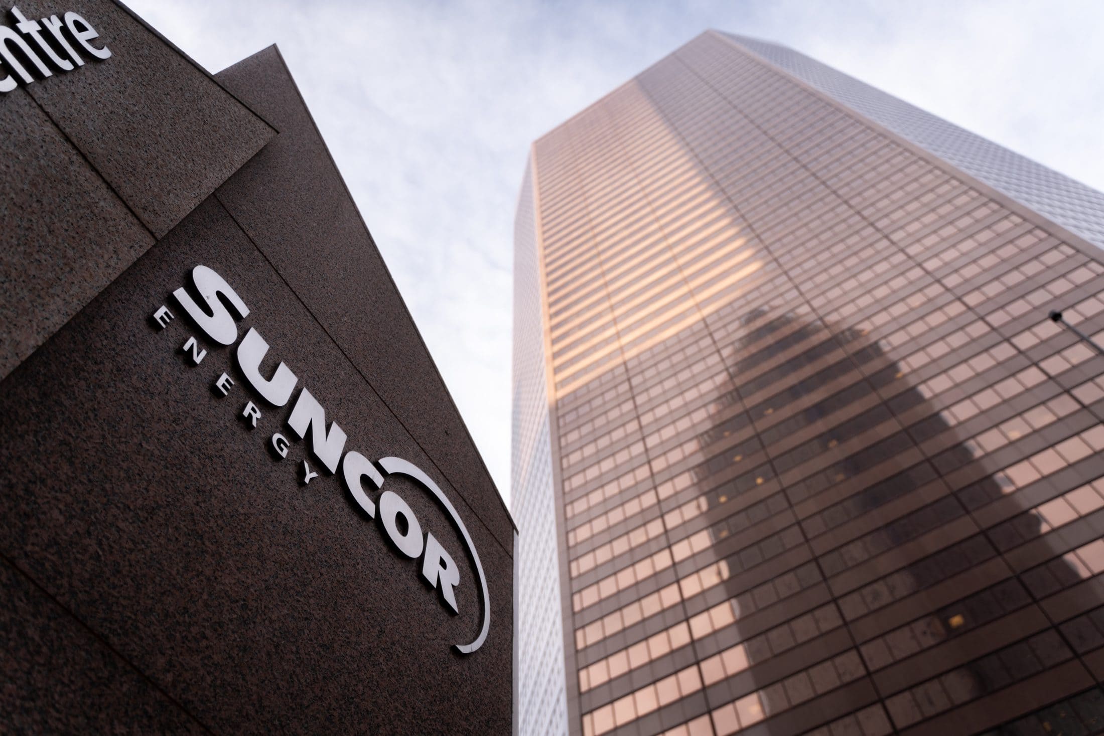 Looking up from the ground at a dark brown sign that says "Suncor Energy" and a light brown office tower in the background.