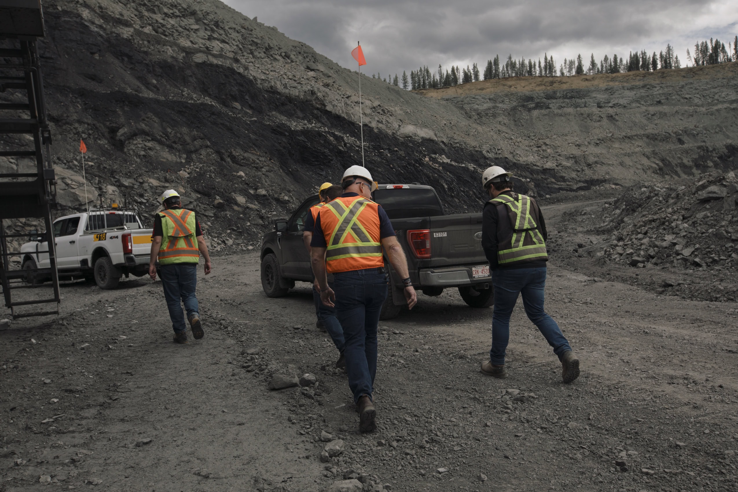 Workers wearing hard hats and reflective vests walk to their trucks next to a coal seam in an Albertan coal mine.