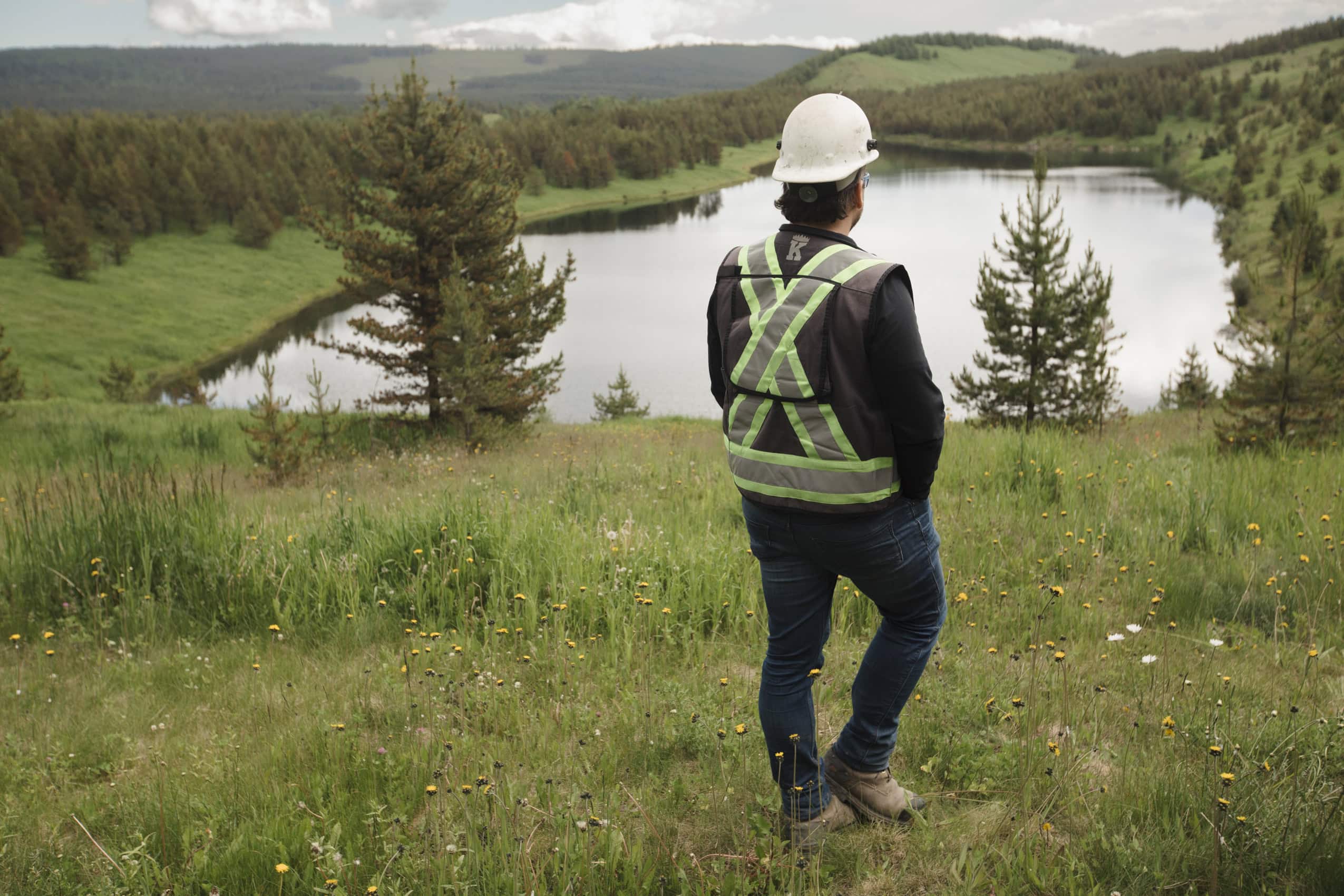 A worker wearing a hard hat and reflective vests overlooks a reclaimed mining pit, which features a large pond surrounded by trees and grasses.