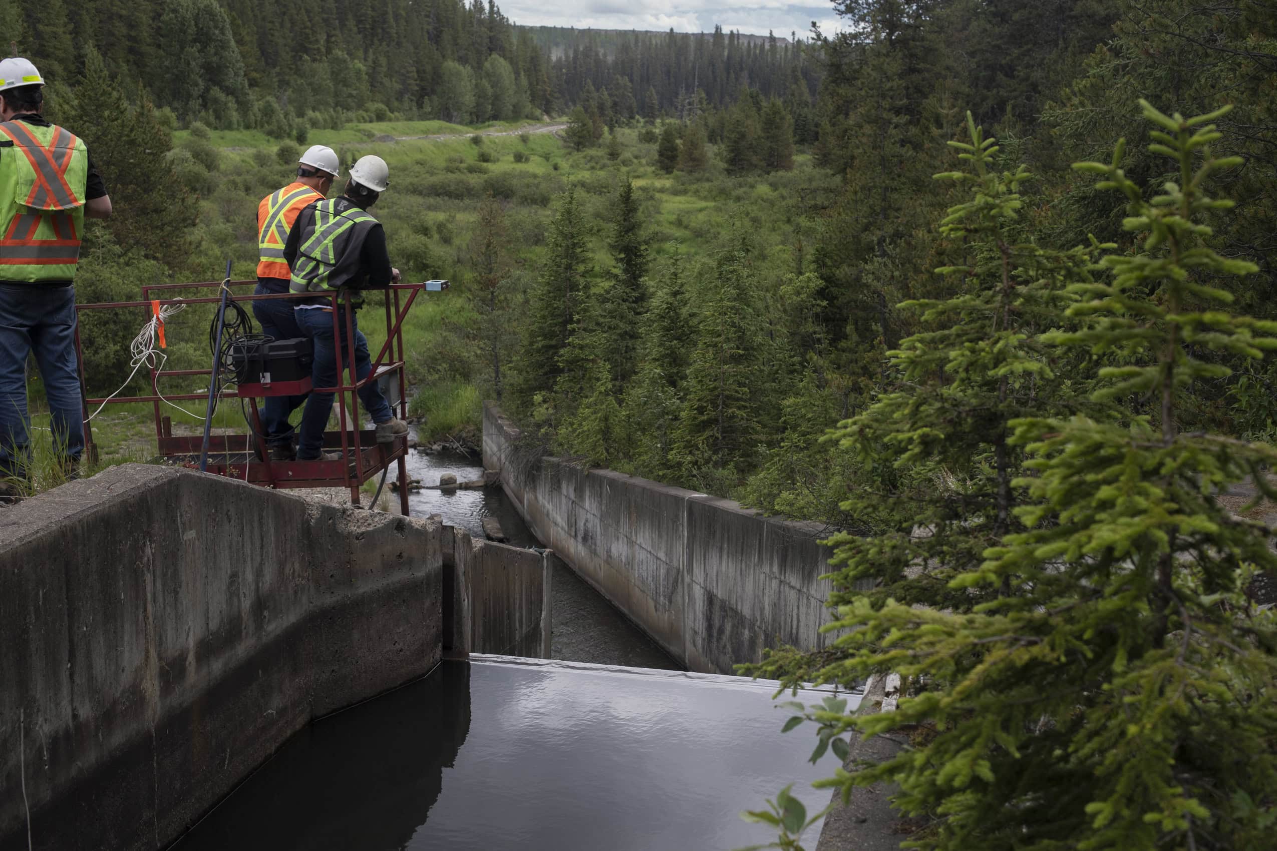 Workers with hard hats and reflective vests overlook a spillway that carriers water out toward a natural landscape beyond.