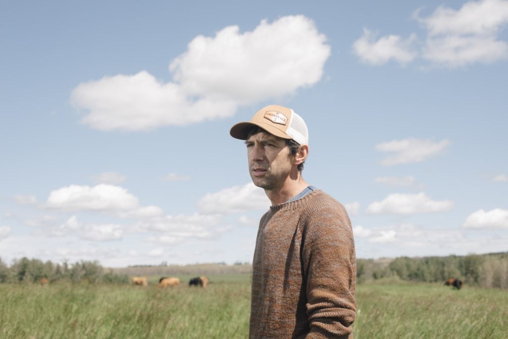 A man in a ballcap and tan sweater standing in a green field, looking off into the distance.