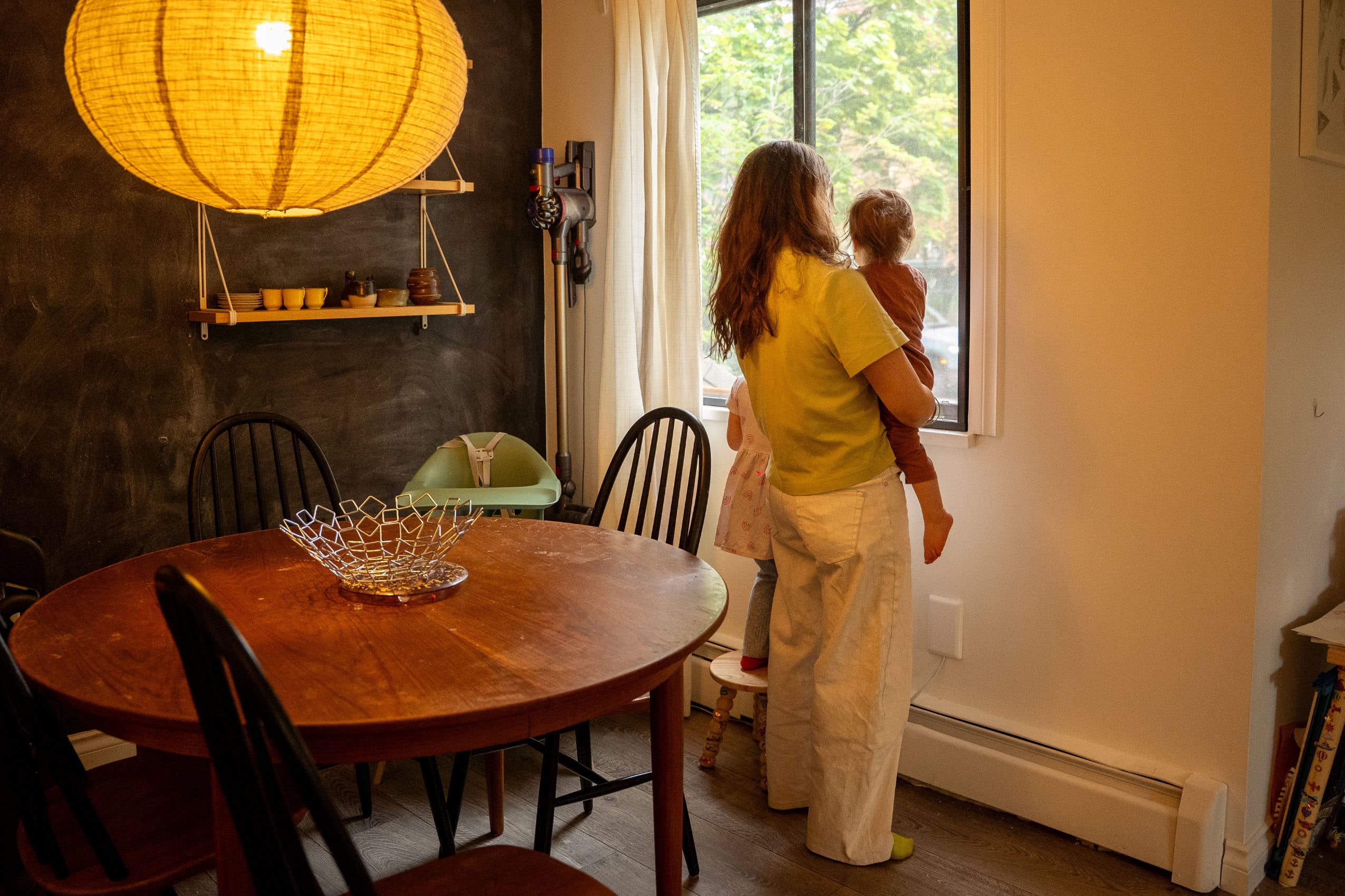 A woman holds her toddler up to their home window to look at the street outside.