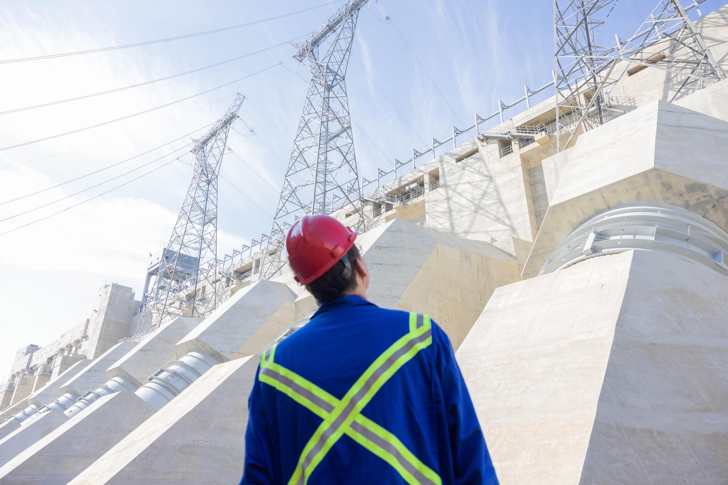 Seen from behind, B.C. Premier David Eby is wearing a hard hat and construction overalls and looking up at the John Horgan Dam near Fort St. John, B.C.