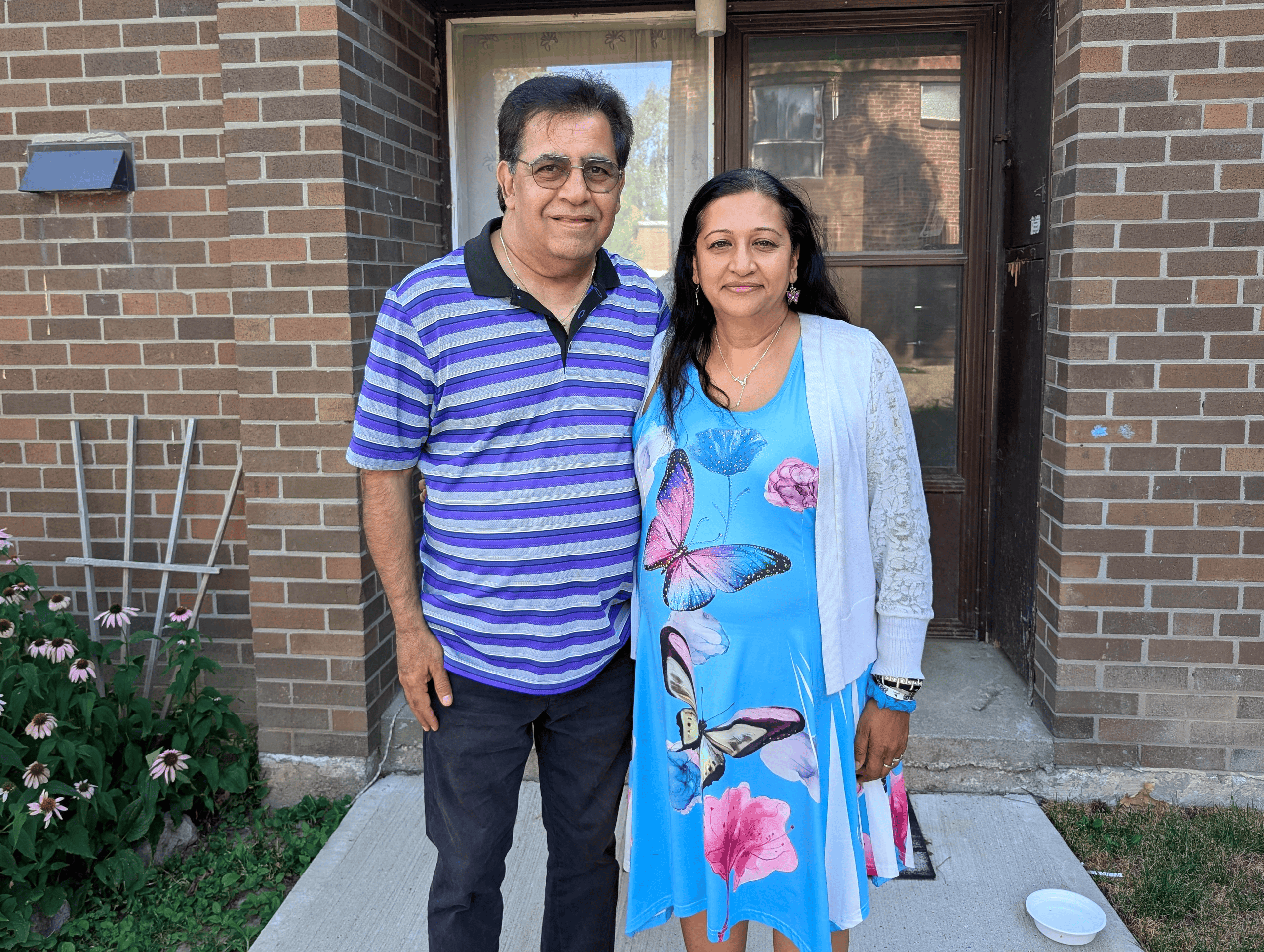 A man and woman stand side by side and smile at the camera in front of their home