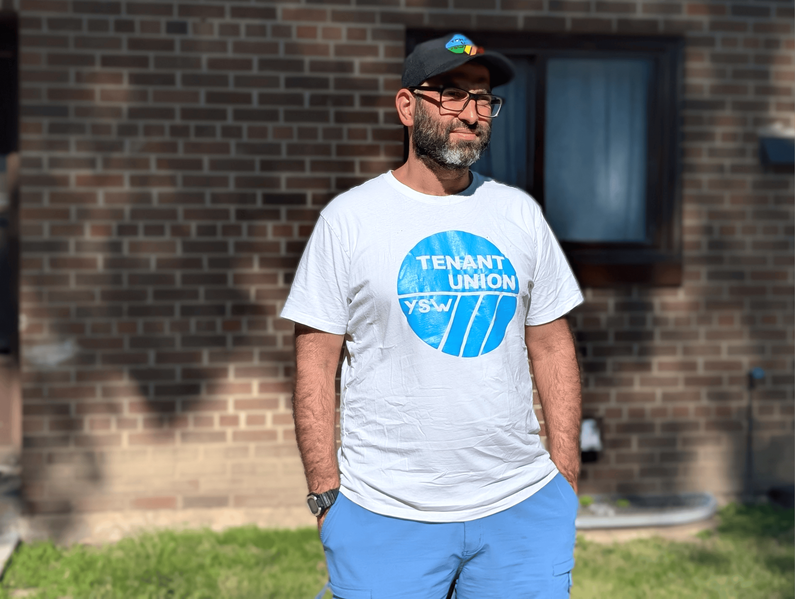 A man with a hat and a shirt that reads "Tenant Union" stands outside a building, he is looking off to the side