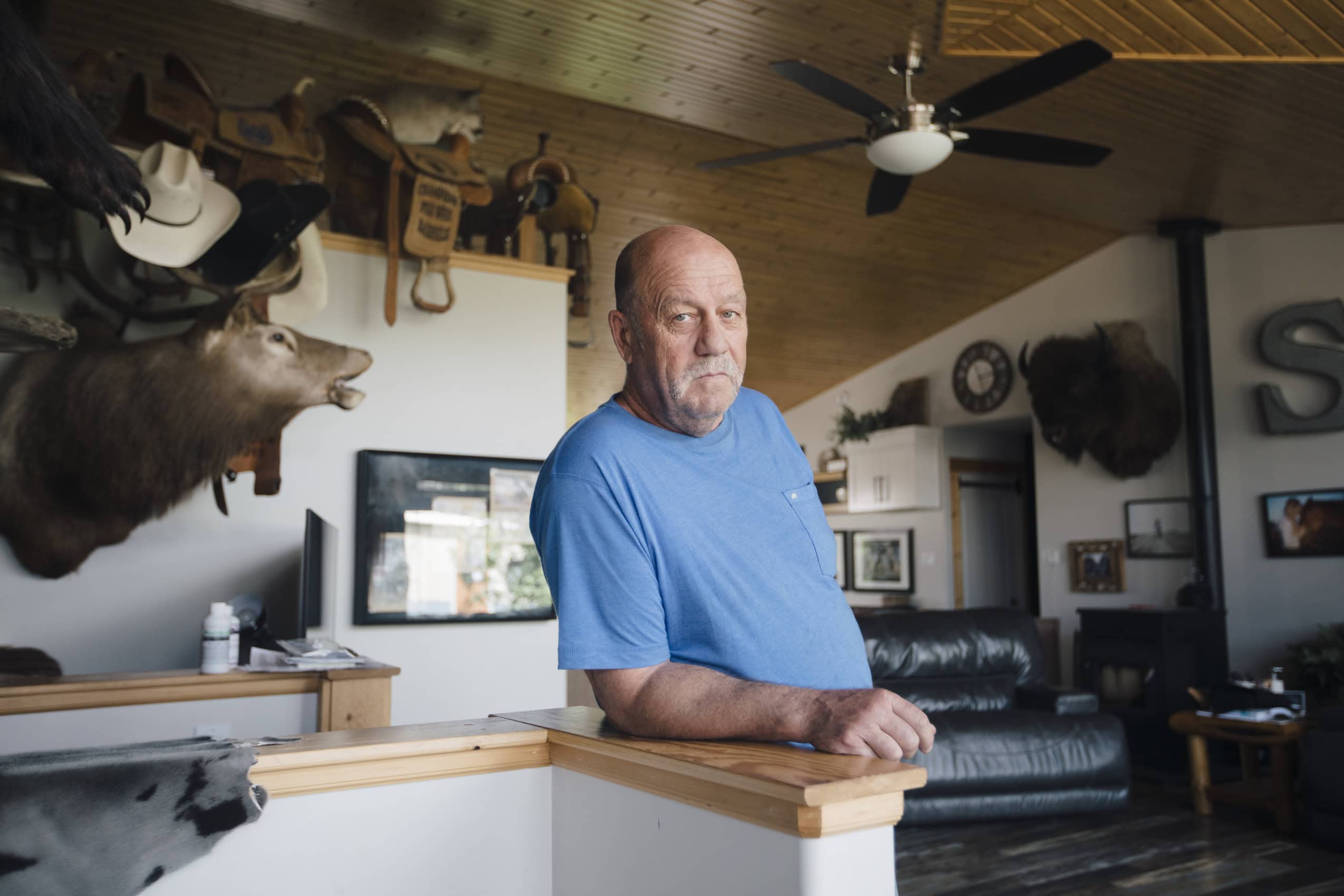A man with a white handlebar moustache leans on a bannister in a home with a stuffed caribou head on the wall.
