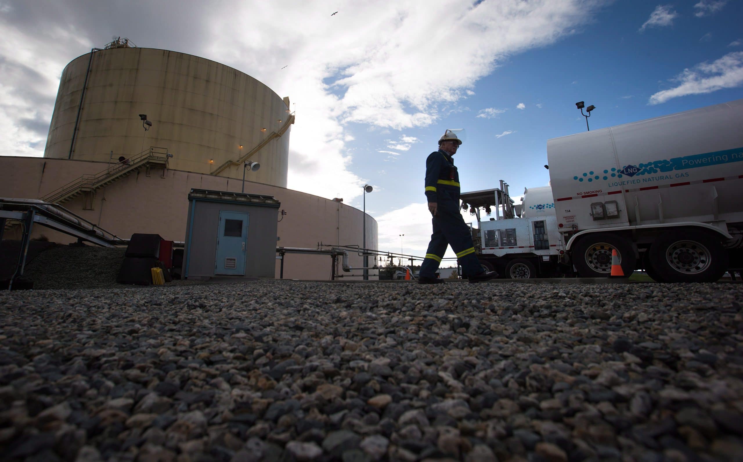 A worker in a blue jumpsuit and white hard hat walks across a gravel lot outside an industrial facility, toward the back of an LNG tanker truck