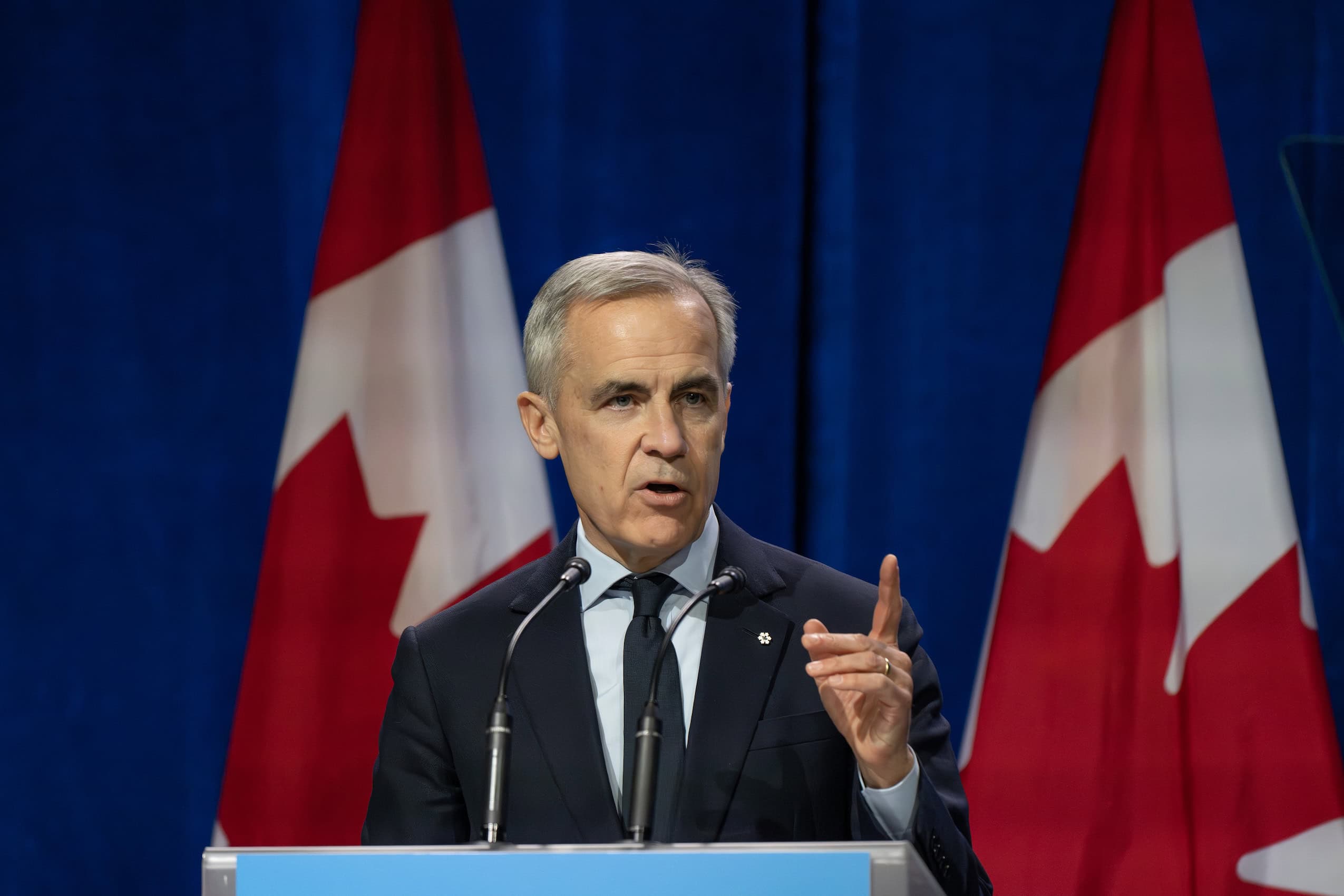 Prime minister Carney, in a dark suit, gestures with his finger while speaking at a lectern flanked by Canadian flags.