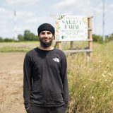 A young man wearing a turban stands in front of a sign that reads Sarbat Farms