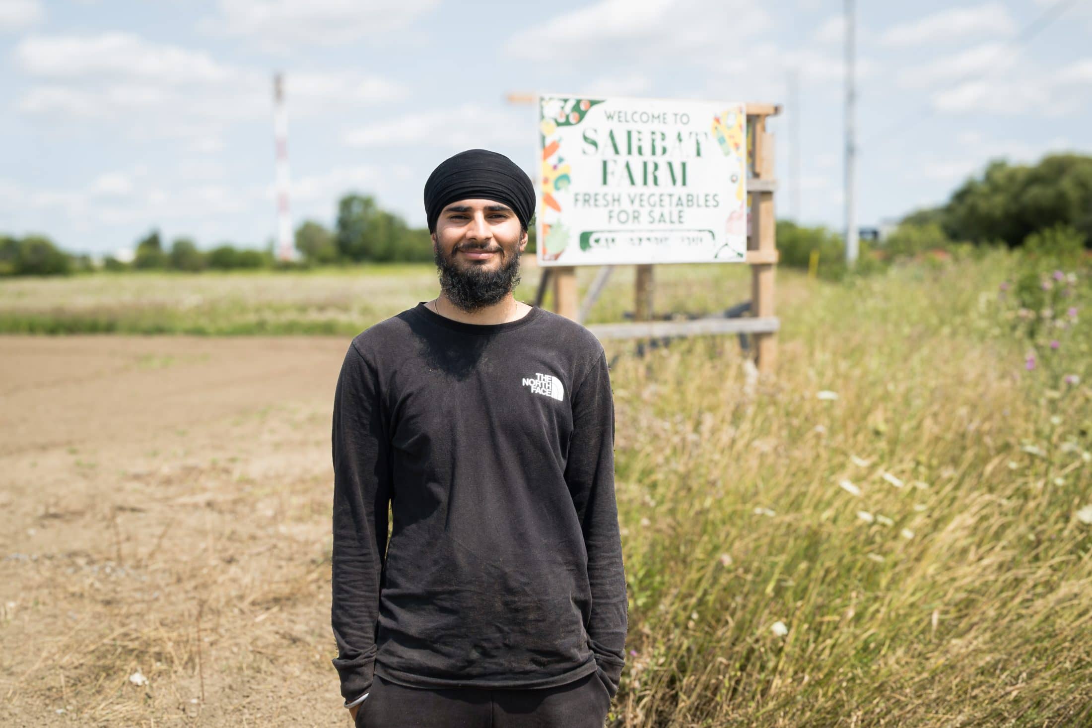 A young man wearing a turban stands in front of a sign that reads Sarbat Farms