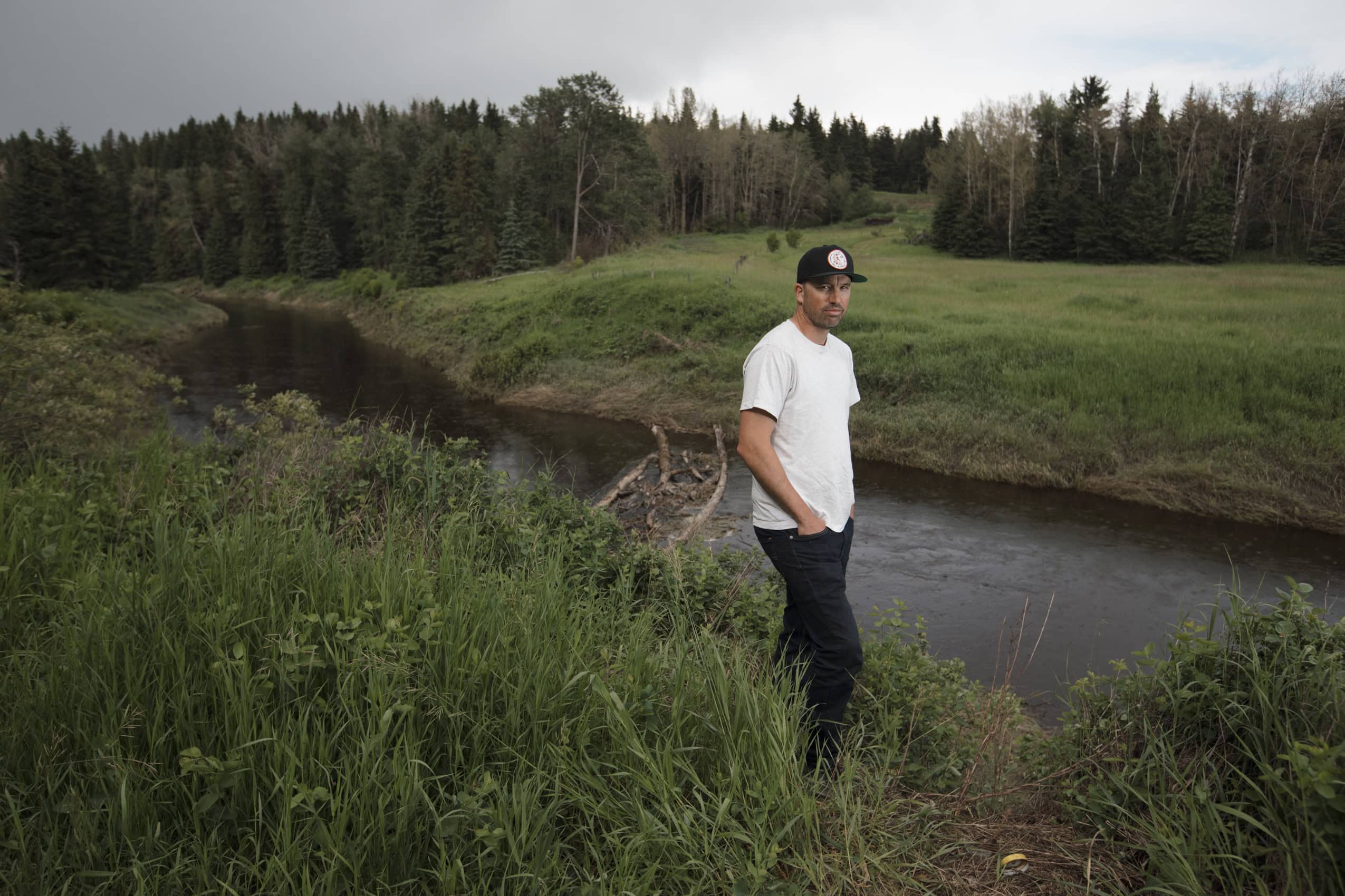 A man in a white shirt and black ballcap stands next to a small river set into a green countryside.