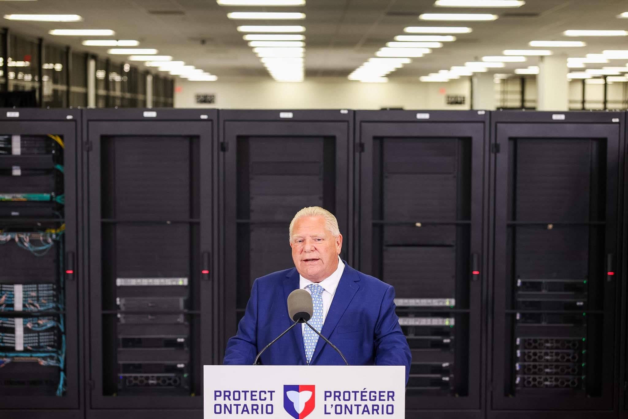 Ontario Premier Doug Ford speaks into a microphone in front of a lectern that reads "Protect Ontario" and "Protéger l'Ontario." Behind him are rows of severs in a data centre.