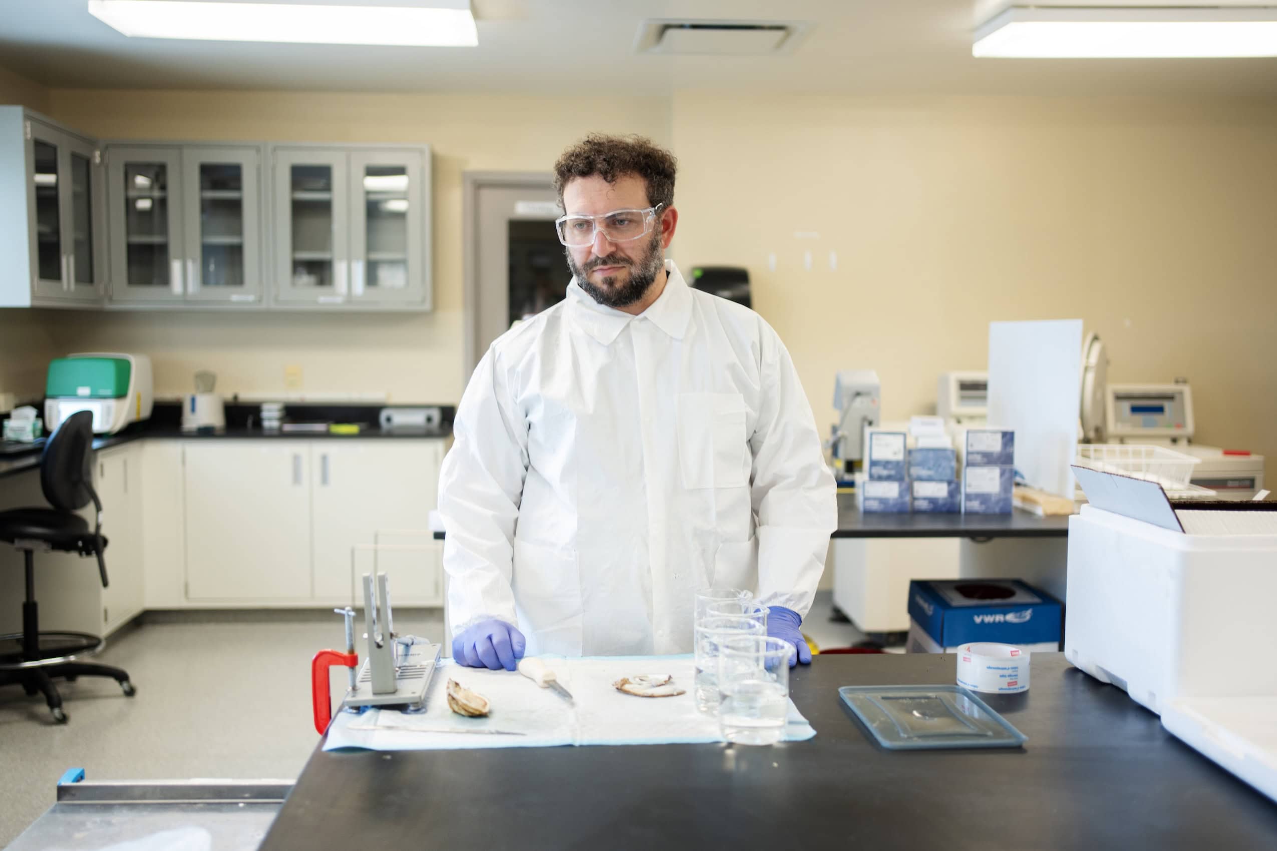 Scientist Tirosh Shapira in a white lab coat and glasses standing at a metal table.