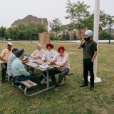 A group of older men sit at a picnic table in a suburban park, while a younger man stands and talks on a cell phone.