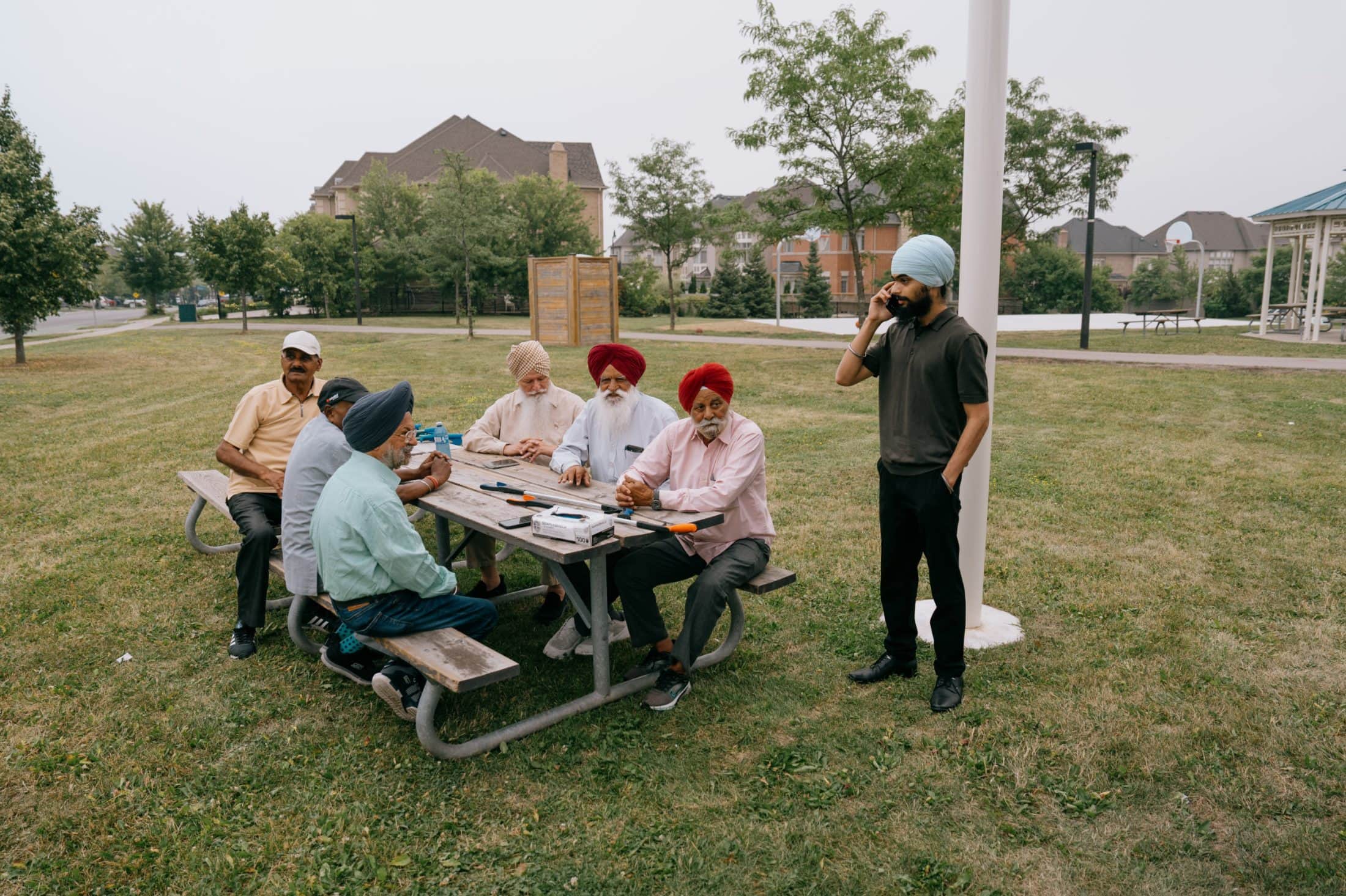 A group of older men sit at a picnic table in a suburban park, while a younger man stands and talks on a cell phone.