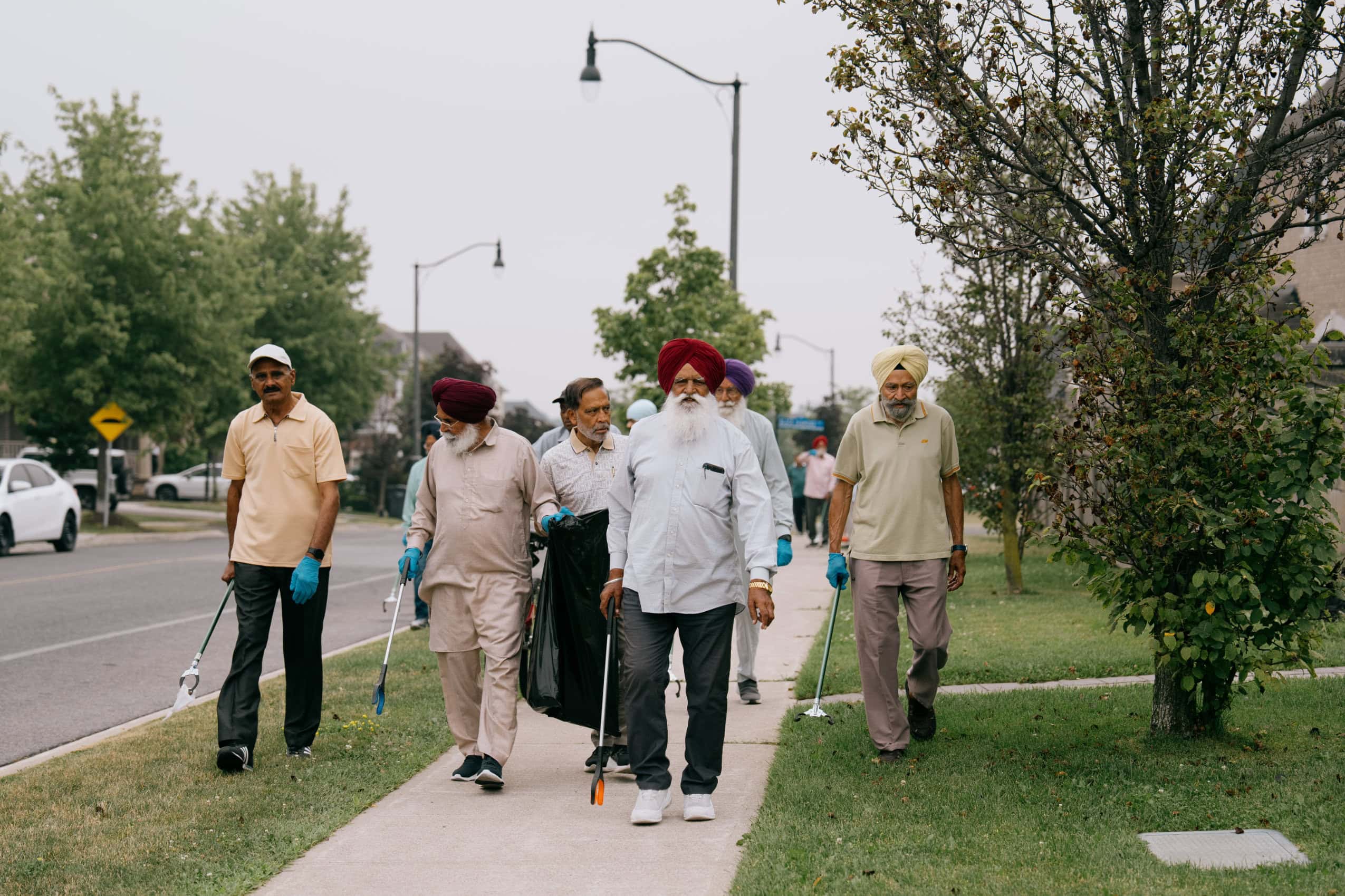 A group of seniors walk along the sidewalk in a suburban neighbourhood, holding litter pickers.