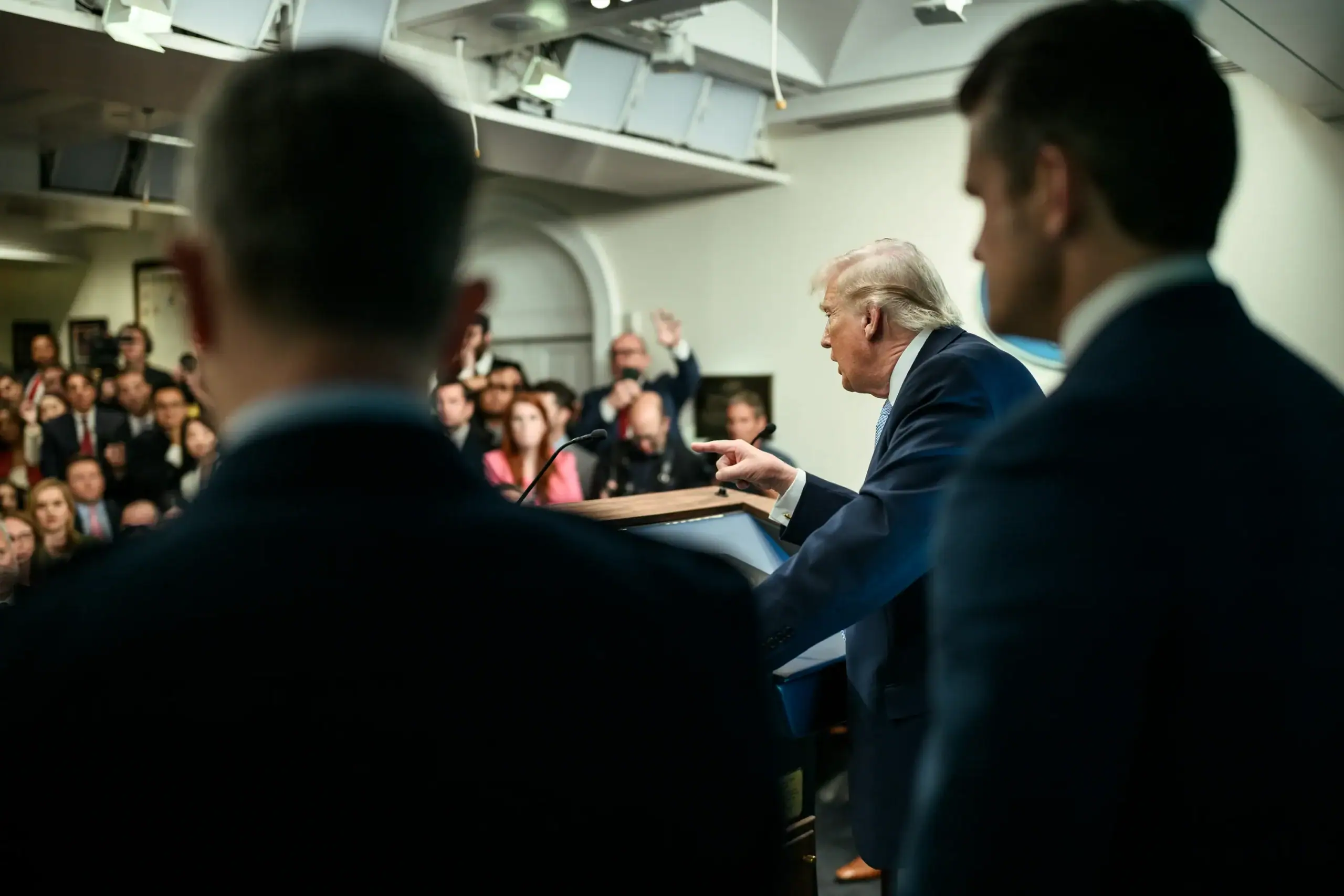Photo from behind of U.S. President Trump gesturing with a finger towards reporters in a windowless room.