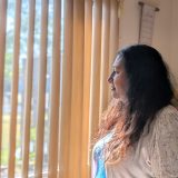 A woman is standing inside her home and looking outside a window