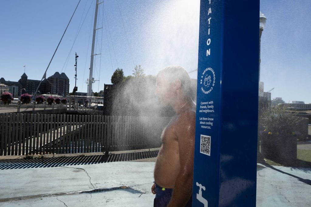 A man stands under a blue misting station, shirtless, mist obscuring his face. Part of Victoria's inner harbour is in the background