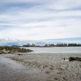 A rocky beach shore along a riverbank that looks out to buildings and clouds and sky.